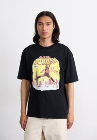 T-shirt en coton noir avec un imprimé graphique coloré d'un homme sautant au-dessus d'une ligne d'horizon de ville, avec "AIR JORDAN" inscrit en lettres majuscules jaunes.