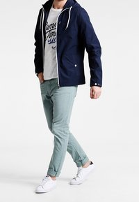 Granatowa kurtka z kapturem i białymi sznurkami, jasnozielone wąskie spodnie oraz białe tenisówki z czarnymi detalami. Widoczna teksturowana tkanina.