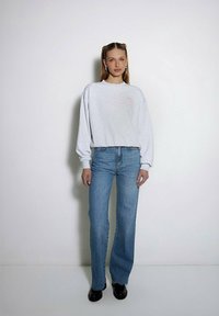 Jasnoszara bluza z długim rękawem, zestawiona z niebieskimi spodniami o wysokim stanie i rozszerzanymi nogawkami. Czarne buty dopełniają outfit. Dyskretne haftowane detale na bluzie.