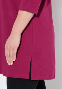 Fuksia tunika lyhyillä hihoilla ja sivuhalkioilla, valmistettu pehmeästä kankaasta. Yhdistetty mustiin legginsseihin, joissa korostuu rento muotoilu.