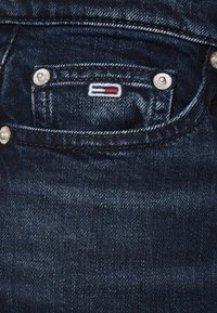 Jeans de mezclilla oscura con un pequeño bolsillo frontal con herrajes plateados y un logotipo bordado colorido cerca del borde del bolsillo.