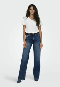 Haut blanc à manches courtes avec décolleté en V, associé à un jean en denim bleu foncé à jambes larges, mis en valeur par une ceinture noire et des chaussures à talons.