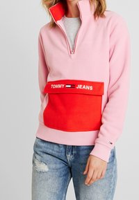 Różowy polarowy sweter z czerwoną kieszenią z przodu, na której jest haft "TOMMY JEANS". Wysoki kołnierz z zapięciem na pół zamka. Noszony z zniszczonymi niebieskimi dżinsami.