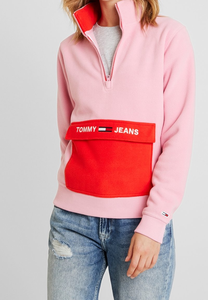 Różowy polarowy sweter z czerwoną kieszenią z przodu, na której jest haft "TOMMY JEANS". Wysoki kołnierz z zapięciem na pół zamka. Noszony z zniszczonymi niebieskimi dżinsami.