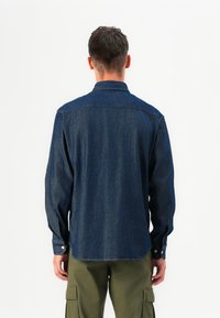 Tummansinisestä denimsärystä, jossa on napillinen kaulus, pitkät hihat ja suora leikkaus. Sisältää hienovaraisia tekstuuri- ja ompeluyksityiskohtia.
