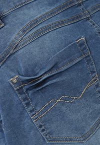 Blå denimtyg med en bakficka som har beige sömnad och ett dekorativt sömmönster. Ytan ser något skrynklig ut.