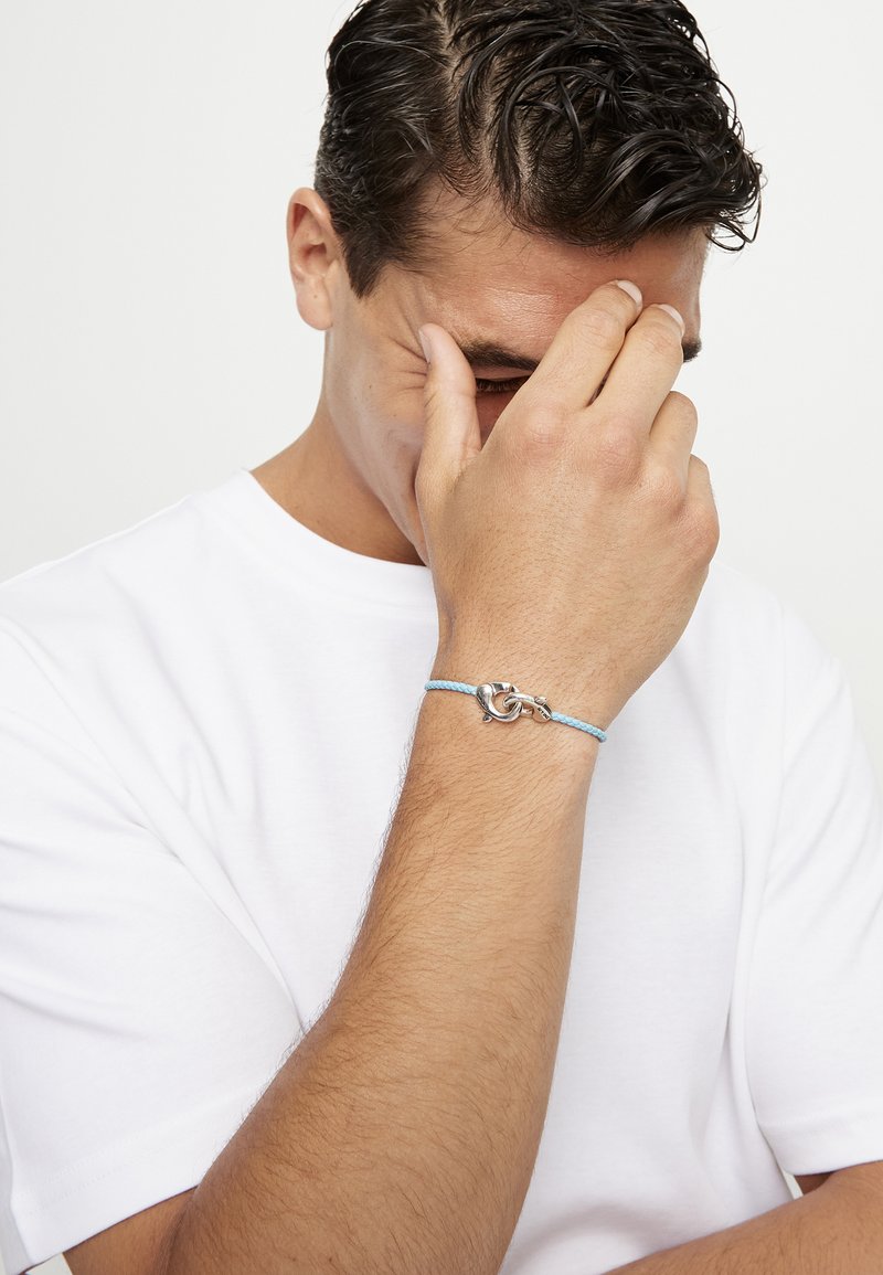 Hombre con camisa blanca tocándose la frente con la mano que lleva una pulsera trenzada azul claro con cierre de plata, sobre fondo blanco.