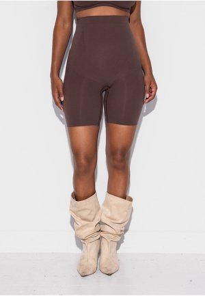 Žena v hnedých biker šortkách s vysokým pásom a voľných béžových semišových čižmách stojaca pred bielou stenou a na bielom podlaží.