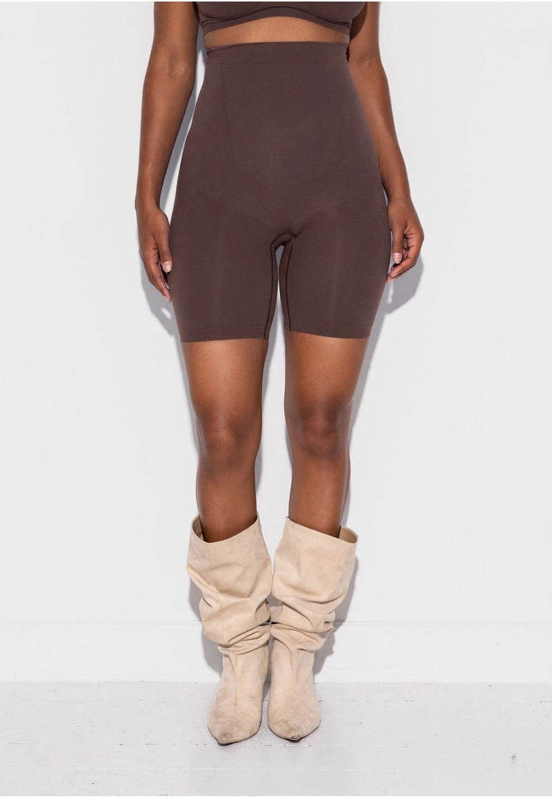 Femme portant un short de cycliste marron taille haute et des bottes en daim beige décontractées, debout devant un mur et un sol blancs.