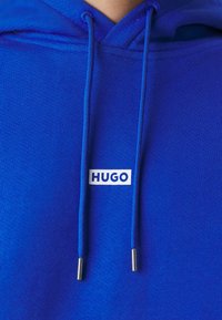 Sweat à capuche bleu avec de longs cordons et un patch rectangulaire blanc centré sous le cou affichant le mot "HUGO" en lettres bleues.