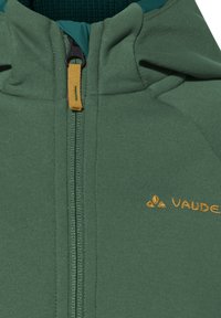 Roheline fliiskamp, millel on ette lukk ja kollane tõmblukk, millel on oranž "Vaude" logo tikand rinnal.