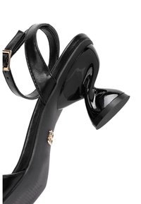 Mustakorkoiset sandaletit, joissa on kiiltävä, kaareva korko, nilkkaremmi ja kultainen koriste remmin lähellä. Näkyvissä on teksturoidun pohjan yksityiskohdat.