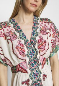 Sukienka haftowana z dekoltem w kształcie litery V, ozdobiona misternymi kwiatowymi wzorami w kolorach różowym, zielonym i niebieskim na jasnym materiale, szczegółowo wykończona guzikami i akcentami.