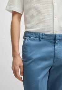 Light blue trousers made of smooth fabric, featuring a flat front, side pockets, and a subtle metallic accent on the waistband.