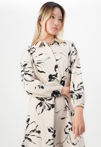 Jeune femme aux longs cheveux blonds portant une robe beige avec un imprimé floral noir, regardant légèrement sur le côté devant un fond blanc.