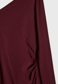 Haut à manches longues bordeaux en tissu doux, avec un détail froncé sur le côté, créant une silhouette ajustée avec une texture lisse.