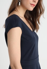Mujer con cabello largo castaño que lleva un vestido azul marino de escote en V y un pendiente de aro plateado con un colgante rectangular.
