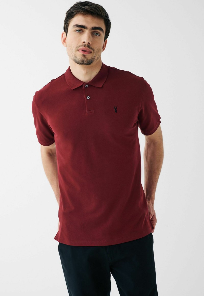 Burgundska polo majica s klasičnom okovratnikom, tri gumba i malim crnim logotipom na prsima. Izrađena od glatkog, prozračnog materijala.