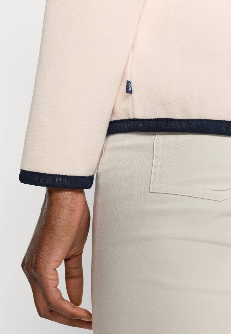 Helles rosa Fleece-Jacke mit marineblauen Akzenten am Saum, mit einem Calvin Klein-Logo. Modell trägt beige Hosen; die Hand ruht auf der Hüfte.