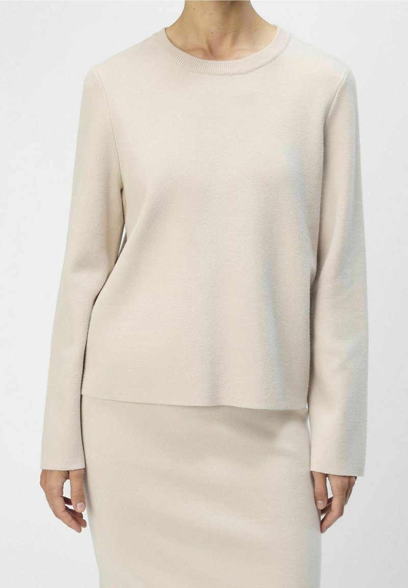 Pull beige à manches longues avec un col rond, texture douce et coupe décontractée, associé à une jupe ajustée assortie.