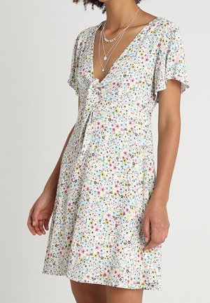 Vrouw draagt een witte jurk met korte mouwen, kleurrijke bloemenprint en een verstelbare strik aan de voorkant, gecombineerd met gelaagde zilveren kettingen.