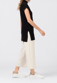 Nainen, joka pitää mustaa hihattomaa neulepaitaa, jossa on sivuleikkaukset, valkoisia leveitä 3/4-housuja ja valkoisia lenkkareita, joissa on beigejä yksityiskohtia, kävelee oikealle.