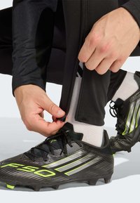 Sorte atletikbukser med lynlåse ved manchetterne, som fastgøres over hvide sokker, suppleret med sorte og neongrønne fodboldsko. Detaljerede teksturer er synlige.