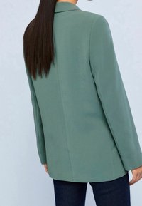 Blazer vert à la coupe structurée, manches longues et texture lisse, avec une couture centrale et des revers ouverts.