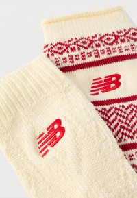 Pletene rukavice v krémovej farbe s červenými akcentmi; jedna má geometrický vzor a druhá dizajn s vrkočovým pletením, obe s červeným logom.