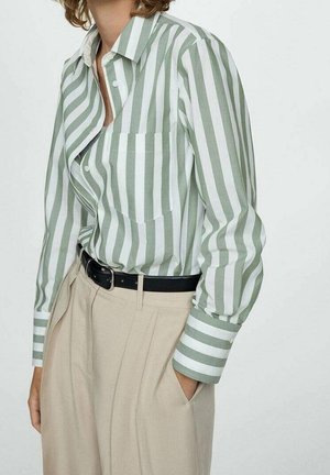Personne portant une chemise rayée verticale verte et blanche rentrée dans un pantalon beige taille haute avec ceinture noire, une main dans la poche.