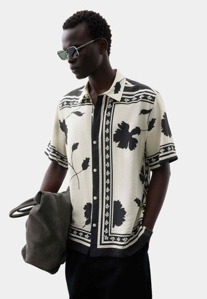 Mann trägt schwarzes und weißes kurzärmeliges Hemd mit Blumenmuster, dunkle Sonnenbrille, hält dunkelgraue Tragetasche, eine Hand in der Tasche.