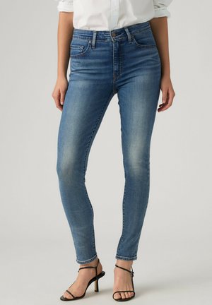 Vrouw draagt strakke blauwe spijkerbroek, een wit overhemd met opgerolde mouwen, en zwarte sandalen met hoge hakken en bandjes, staand tegen een effen achtergrond.