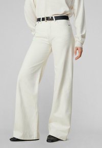 Jeans de pierna ancha en color crema, hechos de una mezcla de algodón, con cintura alta, bolsillos frontales y un cinturón negro con una hebilla dorada.