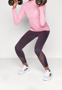 Jasnoróżowa, długorękawowa koszulka sportowa z półzamkiem, zestawiona z ciemnofioletowymi legginsami z panelami z siateczki. Białe trampki z czarnymi akcentami.