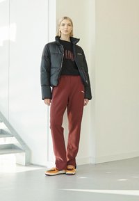 Svart boblejakke, svart genser med tekst, mørkerøde joggebukser og todelte joggesko med oransje detaljer. Minimalistisk innendørs setting.