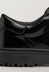 Crna kožna tenisica glatke teksture, zaobljenog vrha, kontrastnih šavova i teksturirane crne gumene potplate s logotipom Adidas.