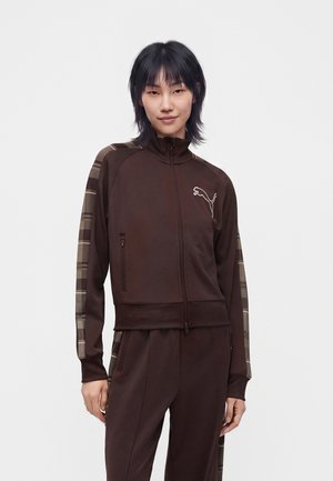 Femme aux cheveux noirs portant un survêtement marron Puma avec des rayures à carreaux sur les manches et le pantalon, sur un fond blanc uni.
