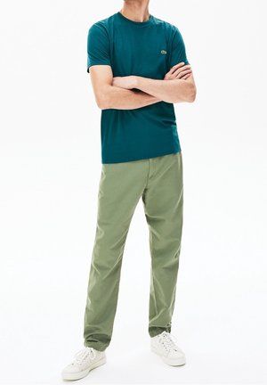 Personne avec les bras croisés, portant un t-shirt à manches courtes bleu sarcelle, un pantalon vert clair et des baskets blanches sur un fond blanc.