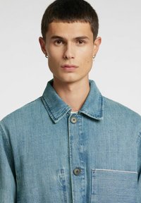 Lichtblauwe spijkerjas met een kraag, twee knopen en een voorzak met contrasterende stiksels. Gestructureerde stof met een ontspannen pasvorm.