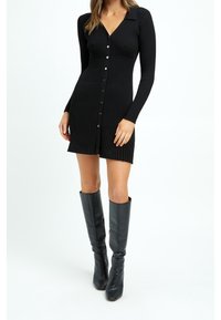 Abito nero in maglia a coste con maniche lunghe, scollo a v e bottoni sul davanti; abbinato a stivali neri in pelle al ginocchio.