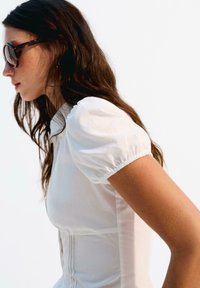 Eine Frau mit langen, dunklen Haaren trägt eine dunkle Sonnenbrille und ein weißes, kurzärmliges Oberteil, während sie vor einem schlichten weißen Hintergrund steht.