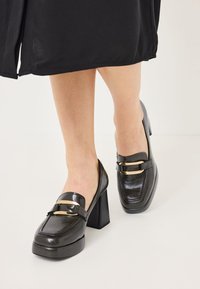 Mocassins à talons carrés en cuir noir avec un bout rectangulaire, accents en métal doré et finition lisse.