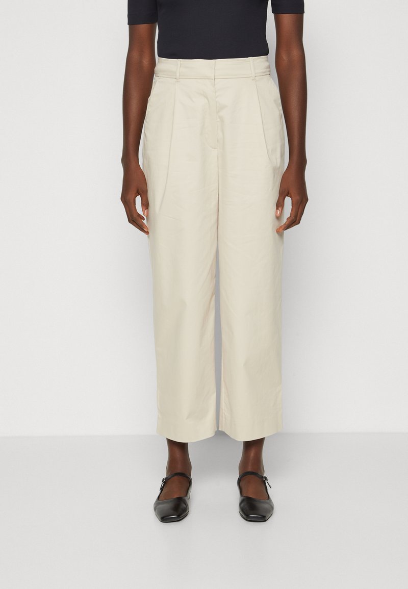 Lichtbeige op maat gemaakte broek met een hoge taille, plooien en een wijde pijpdesign, gemaakt van een zachte stof. Gecombineerd met zwarte platte schoenen.