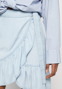 Saia envolvente de jeans azul claro com bainha com babados e laços laterais. Textura suave com costuras visíveis e detalhes de design em camadas.