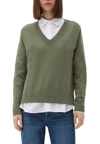 Olijfgroen v-hals sweater van zacht materiaal met geribde manchetten en zoom, gelaagd over een witte blouse, gedragen met blauwe jeans.