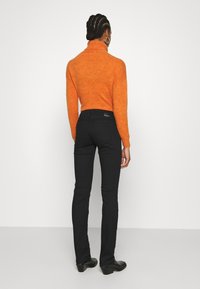 Suéter de cuello alto naranja con un acabado texturizado, combinado con jeans negros ajustados. El atuendo se complementa con botas de tacón negro hasta el tobillo.