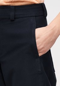 Main insérée dans la poche avant d'un pantalon bleu marine foncé avec des coutures visibles et une passants de ceinture, porté avec un haut marron clair.