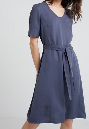 Kobieta ubrana w niebieską sukienkę do kolan z krótkim rękawem, dekoltem w serek oraz paskiem z tego samego materiału przewiązanym w talii.