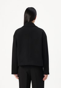 Black cropped jacket with a notched collar, solid fabric, and long sleeves, featuring a straight back seam and minimalistic design.