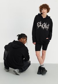 Zwarte hoodies met een opvallende zilveren grafische print, gecombineerd met zwarte shorts en sneakers, op een effen achtergrond. Casual, relaxed model.